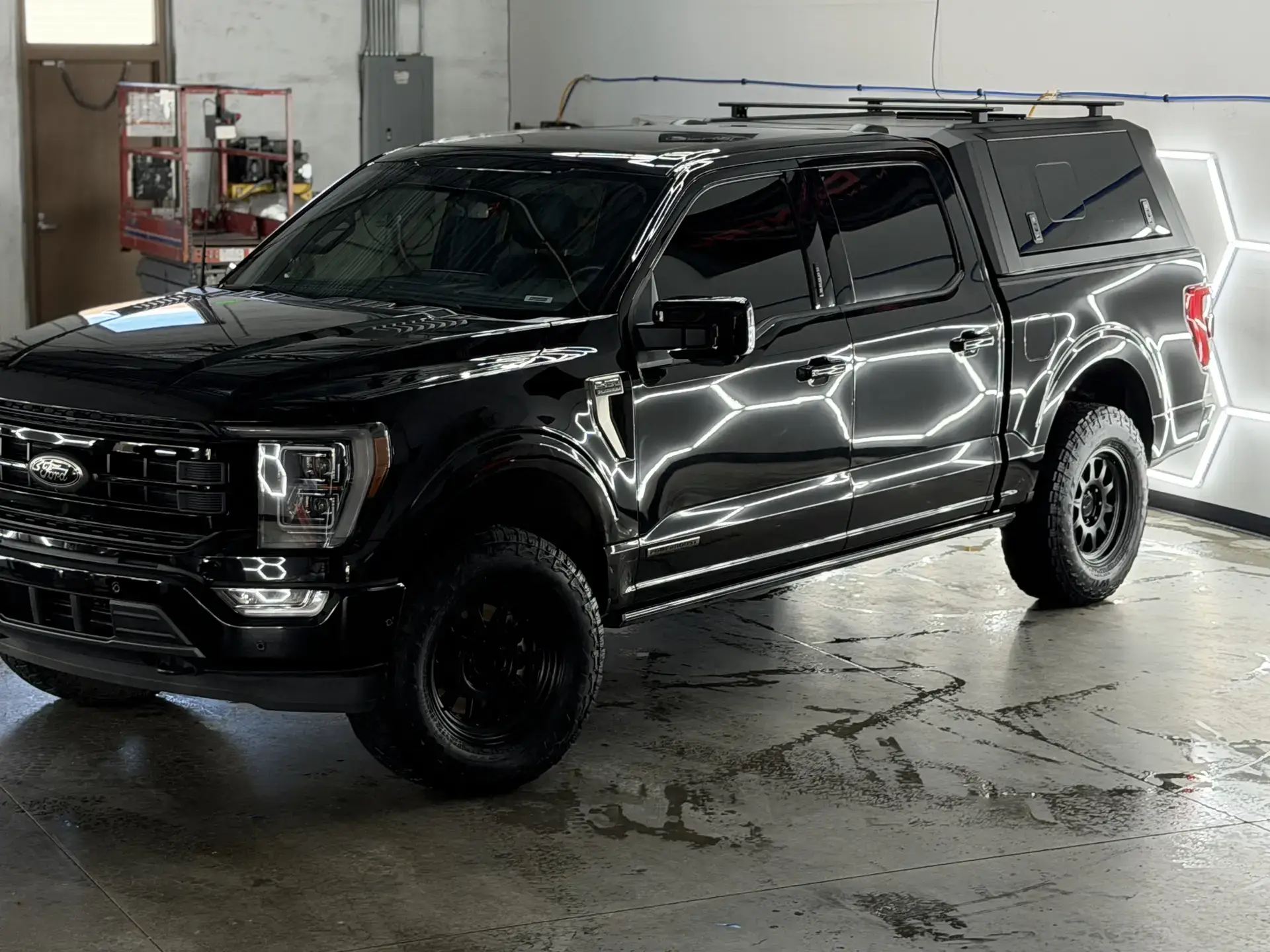 A black Ford F-150 with a camper shell under the studio's hexagon lights, its paint reflecting the light bars