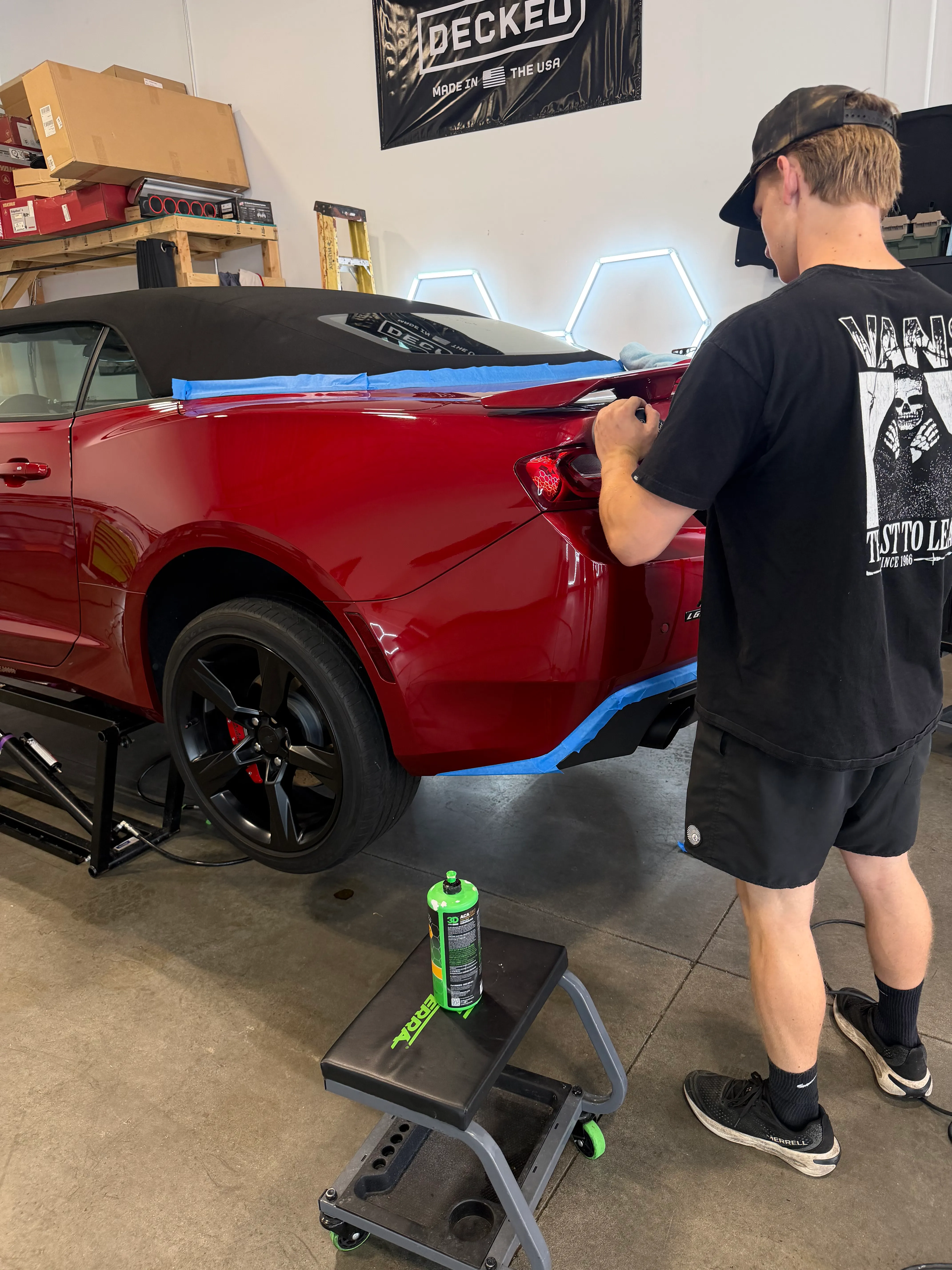 A technician wiping down the taped-off rear quarter of a red Chevrolet Camaro under the studio's hexagon lights