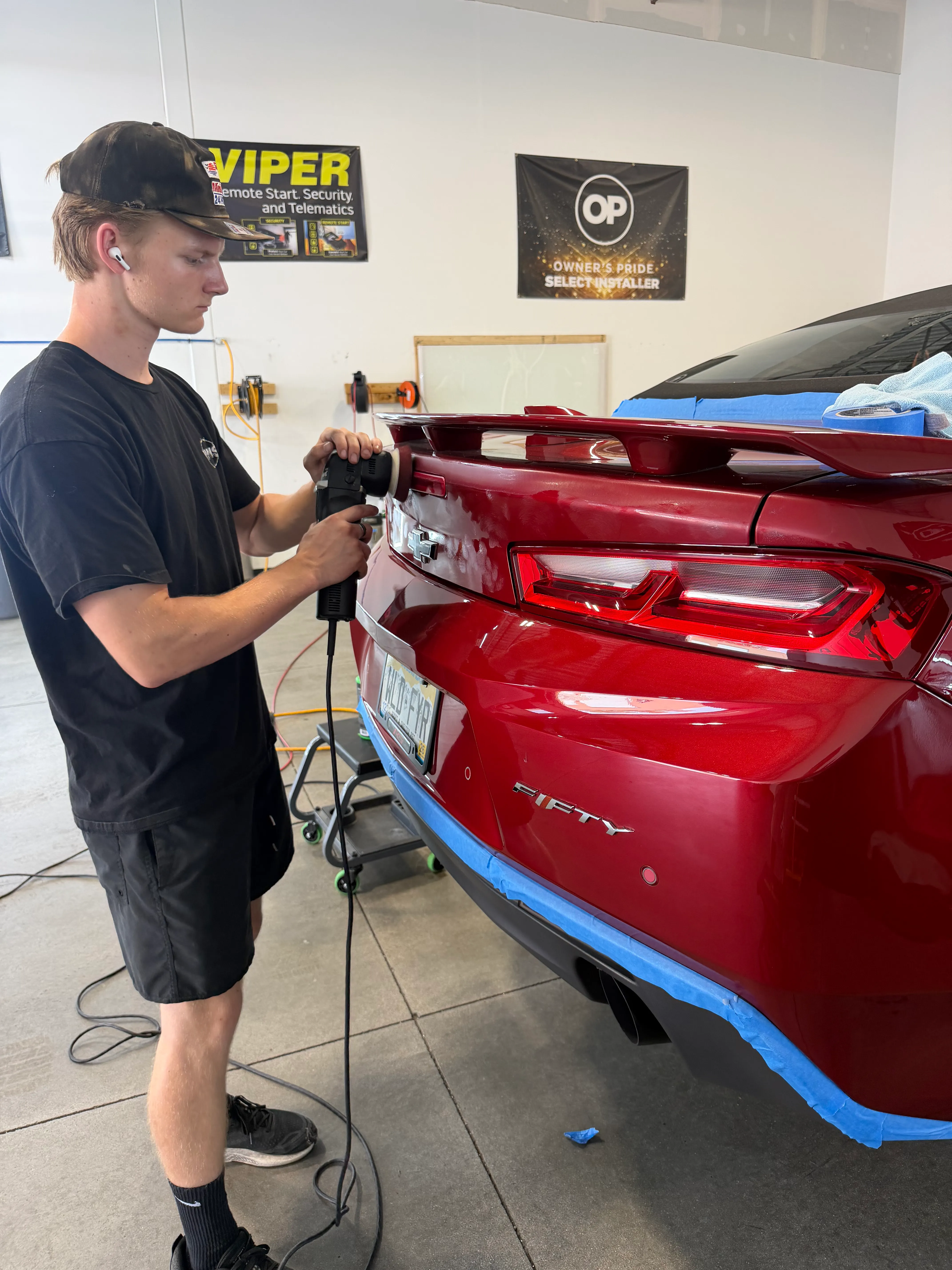 A technician running a dual-action polisher across the taped-off rear quarter of a red Chevrolet Camaro