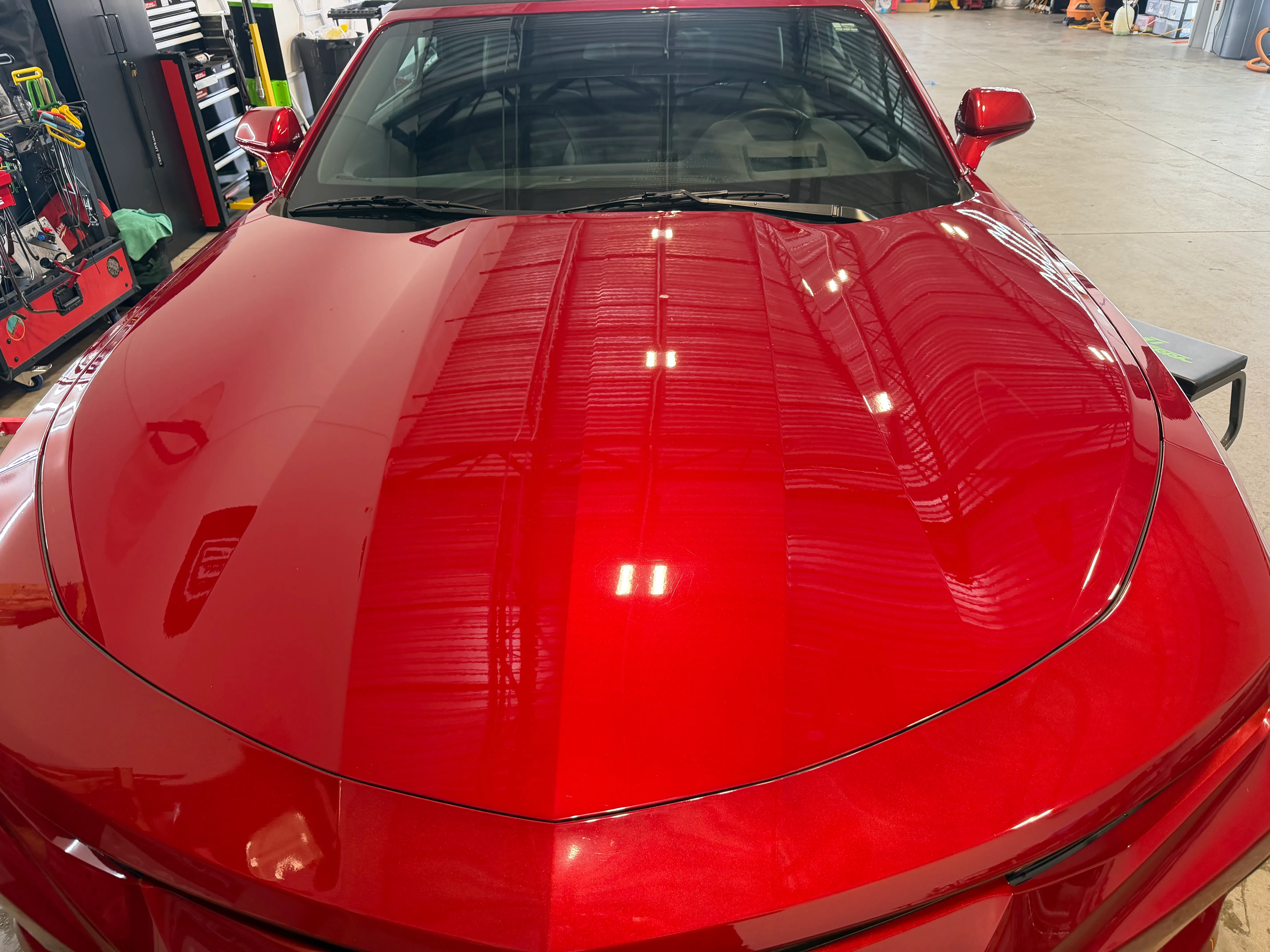The hood of a candy red Chevrolet Camaro reflecting the shop's overhead lights after correction