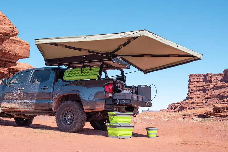 A grey Toyota Tacoma in the desert with a 270 degree awning extended over a tailgate camp kitchen and stacked storage bins