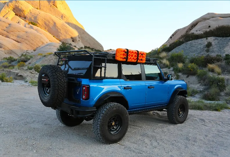 A blue four-door Ford Bronco with a bed rack, orange recovery boards, a rear tire carrier, and fender flares, parked among desert rocks