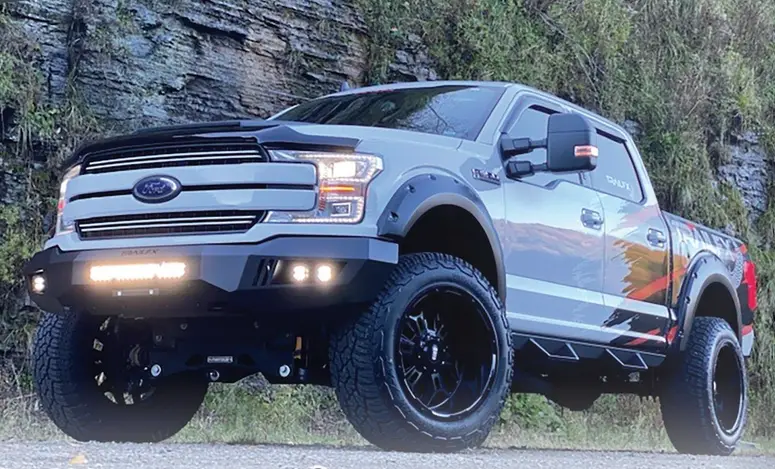 A lifted grey Ford F-150 with a steel front bumper, light bar, fender flares, and black off-road wheels, parked on gravel below a rock face