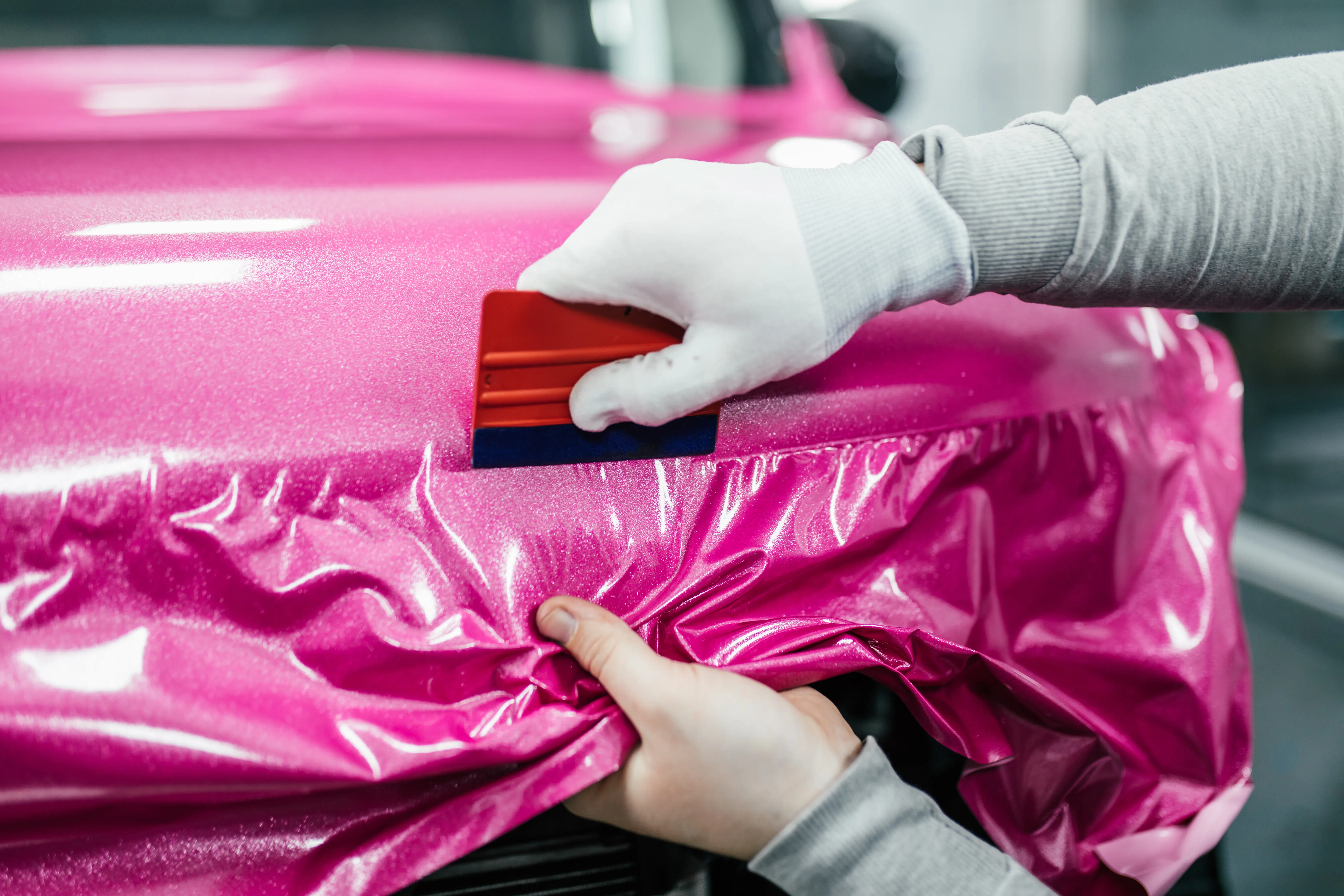 A gloved installer working a squeegee across a hot pink vinyl wrap on a car hood