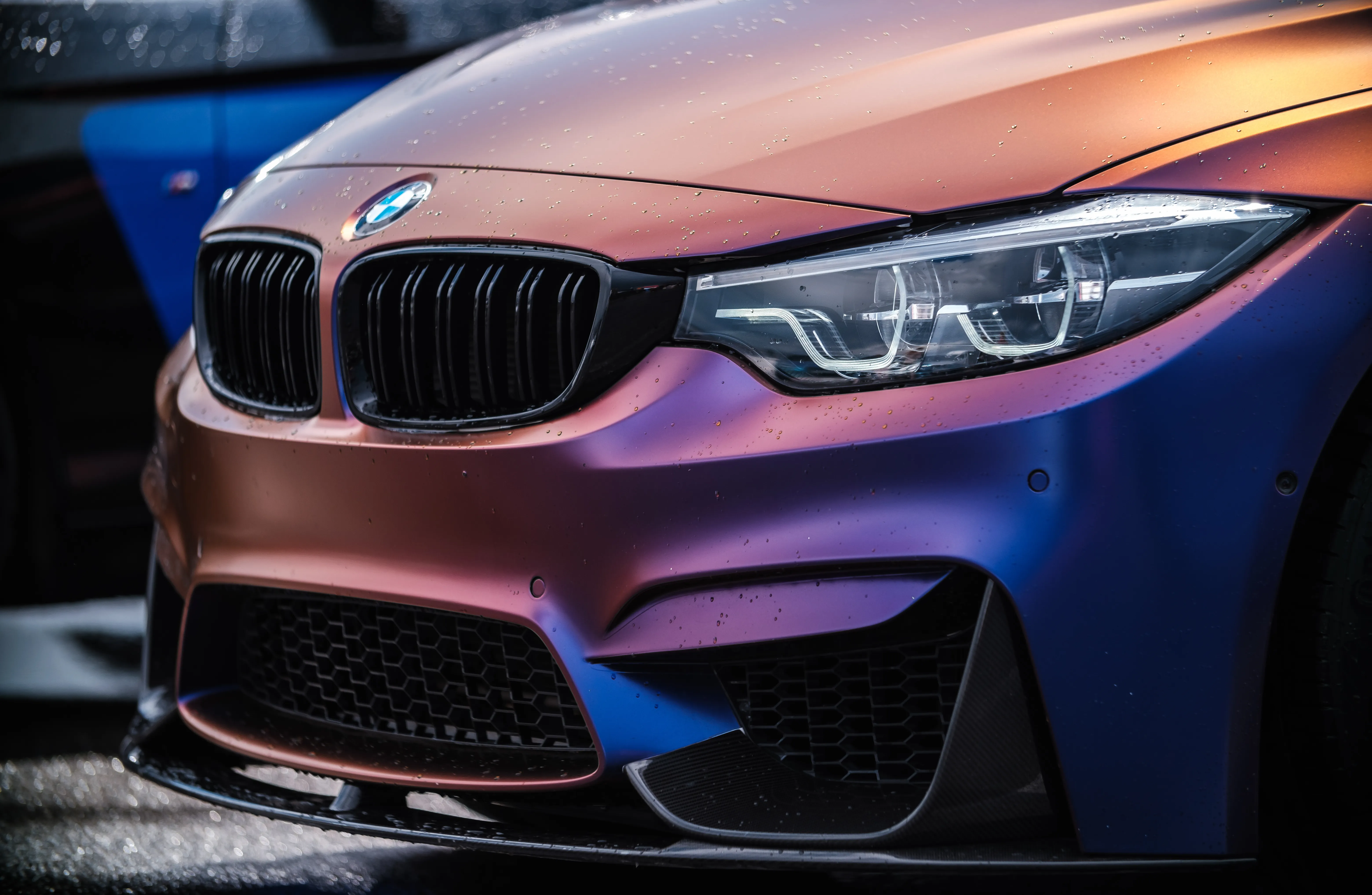 The front end of a BMW M4 in a color-shift wrap that fades from copper through purple to blue, beaded with rain