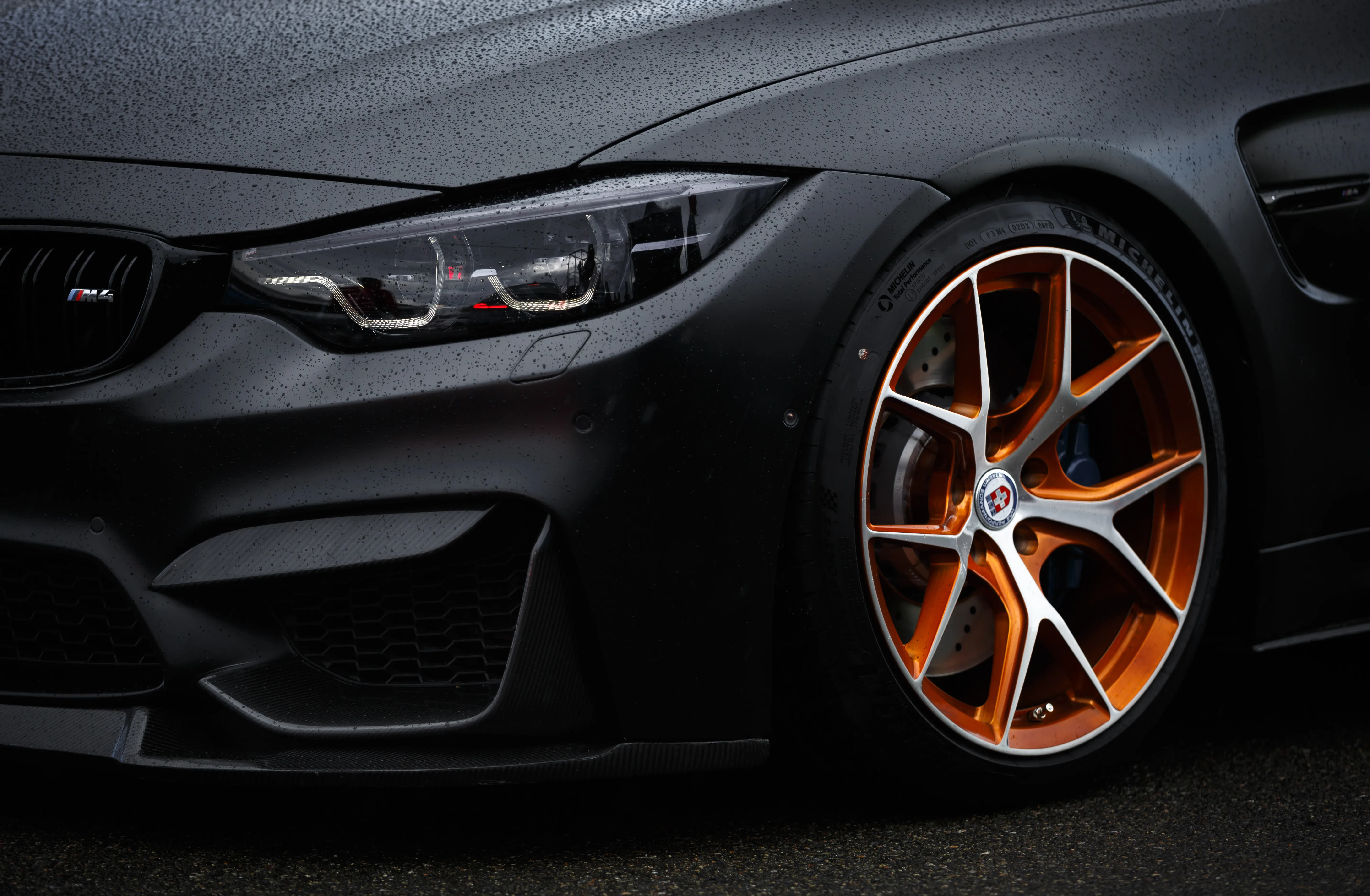 The front corner of a matte black BMW M4 on bronze wheels, wet from rain