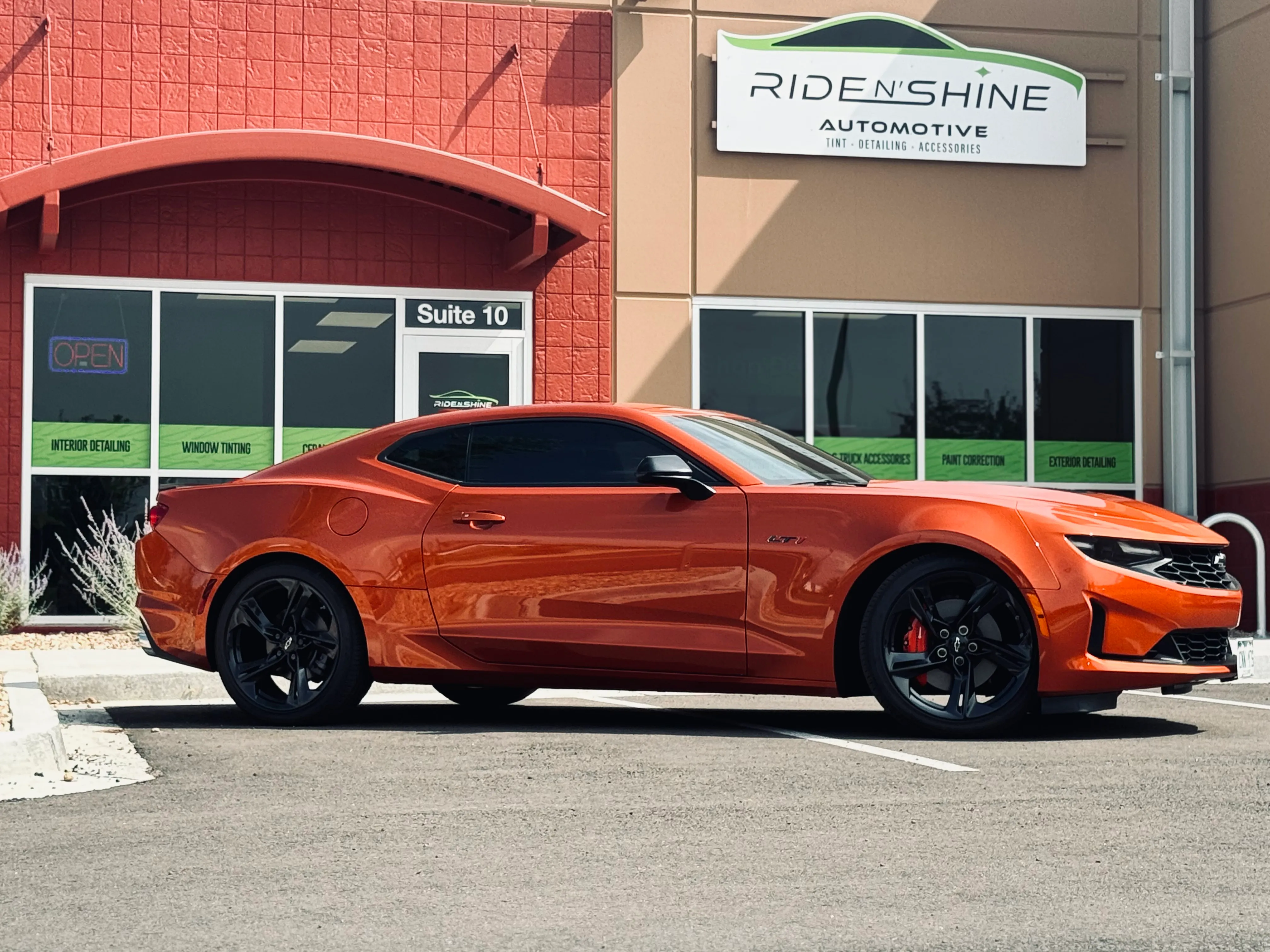 An orange Chevrolet Camaro with tinted windows parked outside the Ride N' Shine storefront